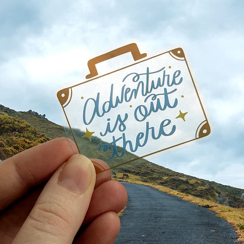 Hand holding a transparent sign with 'Adventure is out there' text against a scenic background.